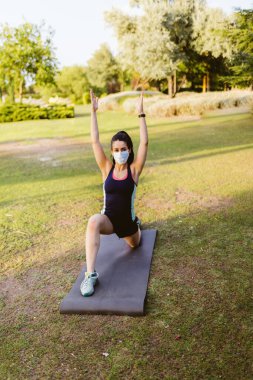 Maskeli genç esmer kadın parkta paspasın üzerinde egzersiz yapıyor ve esniyor.