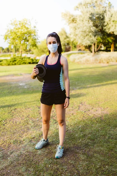 Young brunette female with a mask preparing for exercising and stretching on a mat in the park