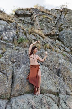 kayaların önünde tahta sopa ile genç beyaz güzel çıplak Amazon kadın durumda.