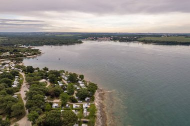 Hırvatistan 'ın Istria kentindeki bir deniz kenarı kamp alanının hava aracı manzarası, turkuaz su, kayalık kıyı şeridi ve yemyeşil ağaçlarla çevrili karavanlar..