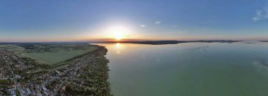 Canlı turuncu ve sarı gökyüzü sakin su yüzeyine yansıyan Balaton Gölü üzerinde nefes kesici bir günbatımı manzarası. Mükemmel altın saat ışıklandırması çarpıcı bir manzara yaratır. Balatonmarafurdo, Macaristan.
