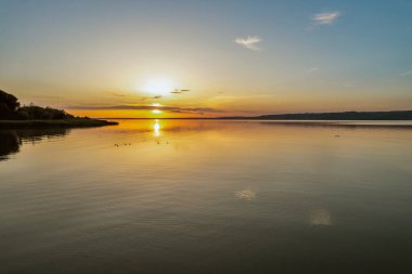 Canlı turuncu ve sarı gökyüzü sakin su yüzeyine yansıyan Balaton Gölü üzerinde nefes kesici bir gün batımı. Mükemmel altın saat ışıklandırması çarpıcı Macar göl manzarası yaratır..