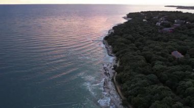 Hırvatistan 'ın Istria kentindeki kayalık bir günbatımı sahil şeridinin hava aracı görüntüsü, kristal berrak turkuaz su, sörf dalgaları ve kayalıkların üzerindeki yemyeşil orman. Arka planda Novigrad.