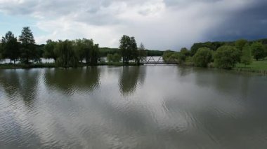 Nagykanizsa yakınlarındaki huzurlu Macaristan kırsalındaki sakin bir ada ve köprüyle birlikte sakin bir tekne gölü üzerinde insansız hava aracı manzarası. Bereketli yeşil bitki örtüsü, ağlayan söğütler cennet gibi deniz manzarası yaratır..