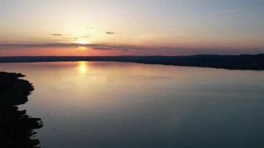 Canlı turuncu ve sarı gökyüzü sakin su yüzeyine yansıyan Balaton Gölü üzerinde nefes kesici bir gün batımı. Mükemmel altın saat ışıklandırması çarpıcı Macar göl manzarası yaratır..