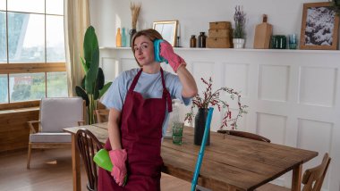 Genç bir kadın odayı lastik eldivenlerle temizler ve kulağının yanındaki fırçayı telefon gibi tutar.