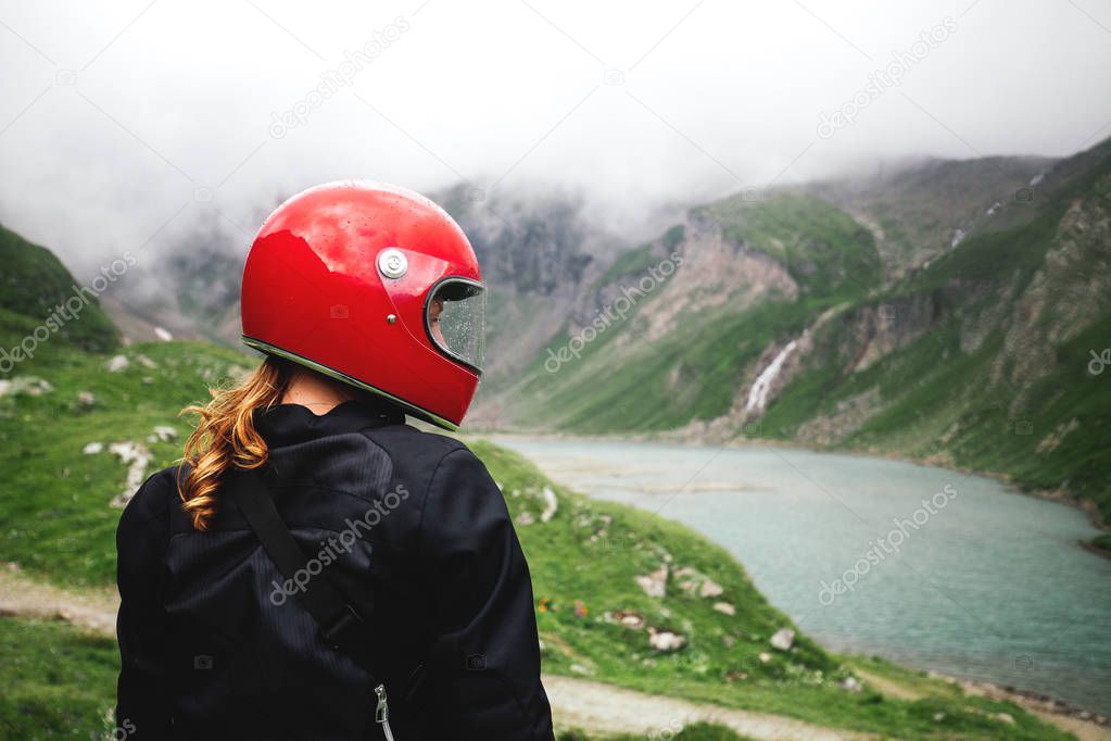 Young giner hair motorcyclist woman close-up in a red glossy stylish retro helmet standing near water while rainy foggy green grass mountains on the backgorund