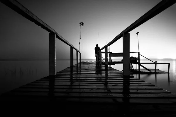 Silueta de un hombre de pie en un muelle de madera en la oscuridad mirando el vacío de un lago con luces brillantes en el lado opuesto — Foto de Stock