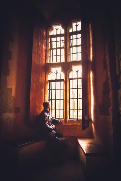 Rear view at silhouette of man sitting in the darkness looking through old bright window with incoming rays of light, dreaming, resting, thinking deeply while waiting quietly and day goes by. — Stock Photo, Image