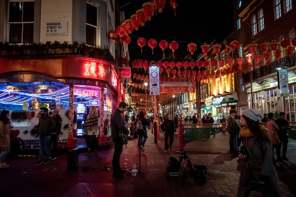 LONDRES, ENGLÂNDIA, 10 DE DEZEMBRO DE 2018: músico de rua tocando guitarra sob luzes de néon em Chinatown, decorado por lanternas chinesas durante o Natal e o Ano Novo, enquanto as pessoas estão passando por Wardour St. . — Fotografia de Stock