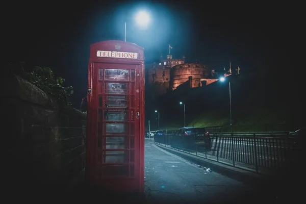 EDINBURGH, SCOTLAND 13 Aralık 2018: Eski İngiliz kırmızı telefon kulübesi gece yol kenarında arka planda şato ile.