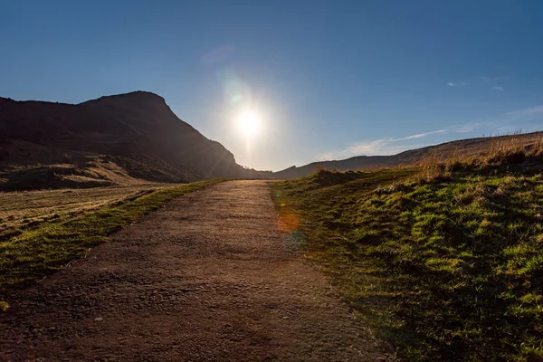 Μονοπάτι κατά μήκος των λόφων του Holyrood Park στο Εδιμβούργο της Σκωτίας, με τον ήλιο να ανατέλλει φωτεινά στο τέλος του δρόμου. Δημοφιλής προορισμός για πεζοπορία και απόλαυση της φύσης. — Φωτογραφία Αρχείου