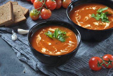 Sıcak kış çorbası. Sarımsak, tatlı kırmızı biber, maydanoz, gri bir arka plan üzerinde iki siyah seramik kase krem ve hamur mayası ekmek ile servis ile kırmızı sıcak domates çorbası 