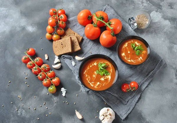 Hot winter soup. Red hot tomato soup with garlic, sweet paprika, parsley, served with cream and sourdough bread in two black ceramic bowls on a gray background