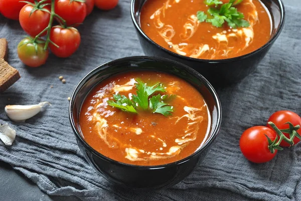 Hot winter soup. Red hot tomato soup with garlic, sweet paprika, parsley, served with cream and sourdough bread in two black ceramic bowls on a gray background