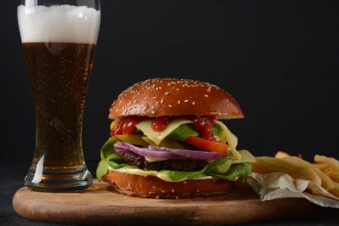 Ev yapımı burger ızgara et, peynir, soğan, domates ve yeşil salata, sos, turşu ve patates kızartması ve arka planda bir bardak bira..