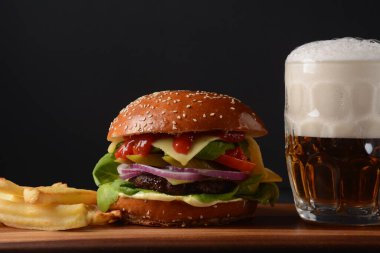 Ev yapımı burger ızgara et, peynir, soğan, domates ve yeşil salata, sos, turşu ve patates kızartması ve arka planda bir bardak bira..
