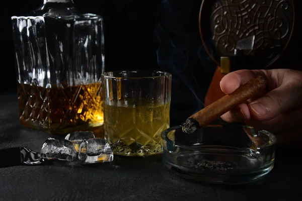 Glass of the whiskey with ice cubes and a square decanter, and cigar on a black  background.