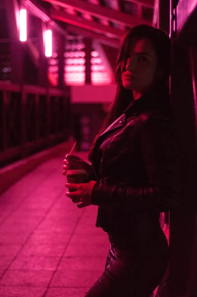 Ein junges Mädchen mit langen schwarzen Haaren in einer schwarzen Jacke mit einem Glas heißen Kaffees in den Händen posiert in einem langen Korridor mit rosa Licht — Stockfoto
