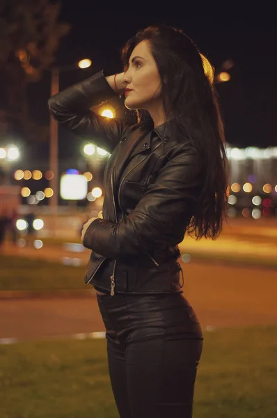 Street style, fashion. Portrait of a woman in black with beautiful makeup and red lipstick on the road with backlight, bright lights and light of lanterns