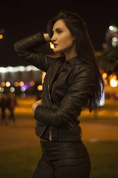 Street style, fashion. Portrait of a woman in black with beautiful makeup and red lipstick on the road with backlight, bright lights and light of lanterns