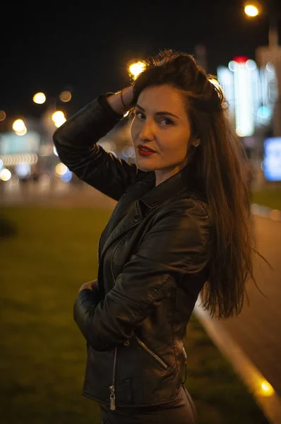 Street style, fashion. Portrait of a woman in black with beautiful makeup and red lipstick on the road with backlight, bright lights and light of lanterns