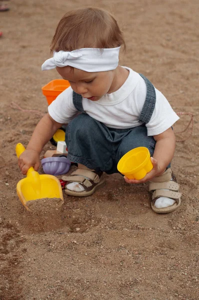 A little girl in white T-shirt, denim overalls, white headband and beige sandals plays in the sandbox with sand. in the hands of a girl shovel and cup. — Stock Photo, Image