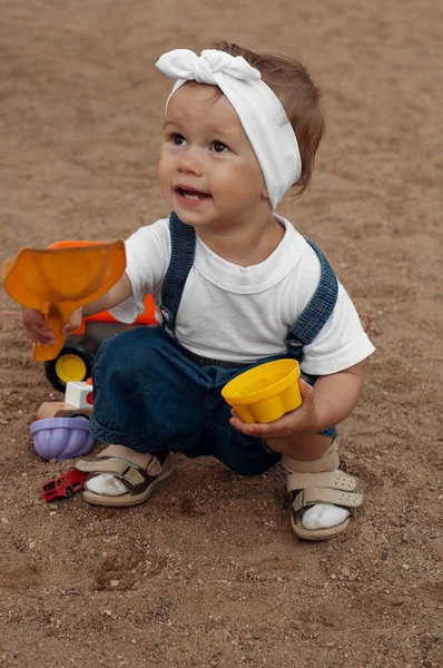 A little girl in a white T-shirt, denim overalls, a white headband and beige sandals sits in a sandbox and plays in the sand. in the hands of a girl shovel and cup. Girl look at mom and smiling. — Stock Photo, Image