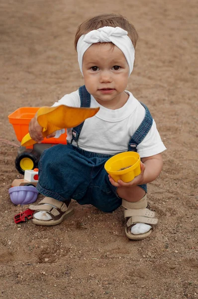 A little girl in a white T-shirt, denim overalls, a white headband plays in the sandbox with sand. in the hands of a girl shovel and cup — Stock Photo, Image