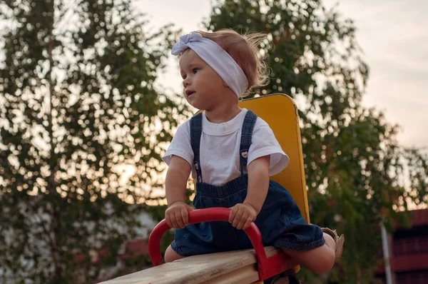 A little girl, 18 months old, in a white T-shirt, denim overalls, white rim and beige sandals sits on a seesaw and looks around — Stock Photo, Image