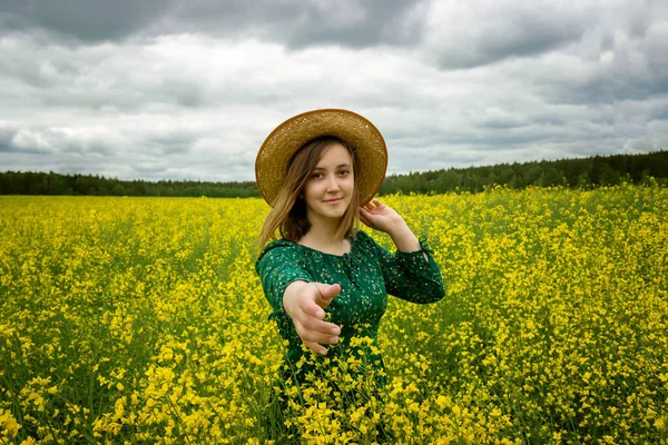 Sarı bir tecavüz sahasında genç, beyaz bir kadın elbisesi ve hasır bir şapka modern bir şapka giyer ve gülümser. El ele tutuşmak konseptimi takip edin. — Stok fotoğraf