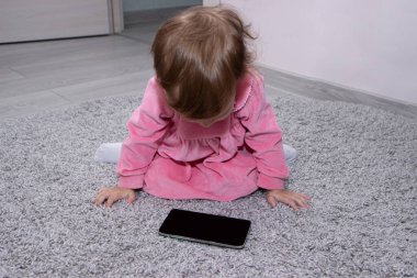 Güzel, küçük bir kız. Odasında halıda akıllı bir telefon var..