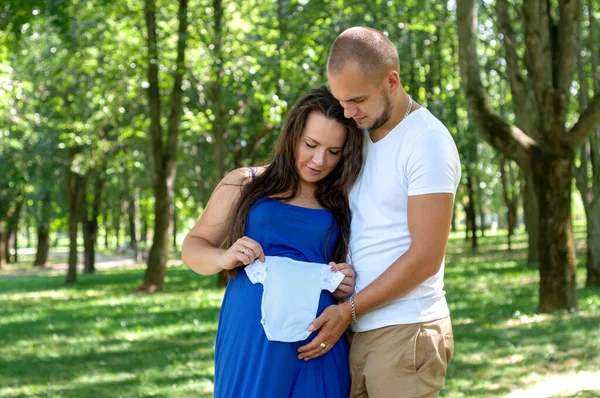 parents-to-be couple pregnancy holding a baby boy blue bodysuit outdoors at green nature background.