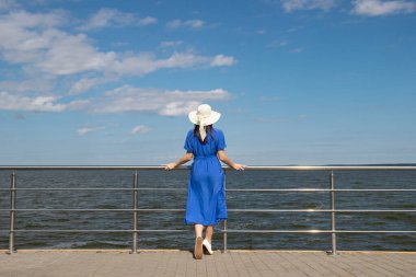 Hasır şapkalı genç kadın zarif elbiseler içindeki iskelede kalıp manzaranın tadını çıkarıyor..