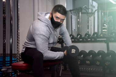 Güçlü sporcu vücut geliştirici, dambıllarla egzersiz yap, koyu renk spor salonunda, tıbbi maske takan sporcu, koronavirüs salgını