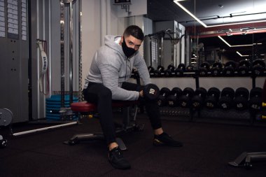 Güçlü sporcu vücut geliştirici, dambıllarla egzersiz yap, koyu renk spor salonunda, tıbbi maske takan sporcu, koronavirüs salgını