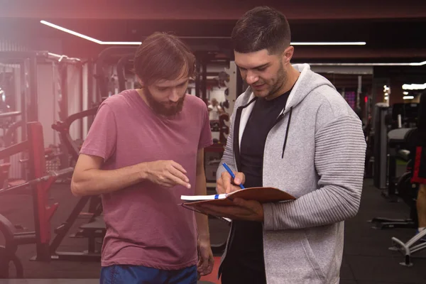 Fitness, sport, exercising and diet concept - smiling young sportsman and personal trainer with clipboard writing exercise plan in gym — Stock Image Fitness Sport Exercising Diet Concept Smiling Young Sportsman Personal Trainer Stock Image