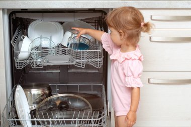 Sevimli sarışın bebek mutfakta yardım ediyor bulaşık makinesinden tabakları çıkarıyor. Çocuk ev işlerine yardım ediyor.