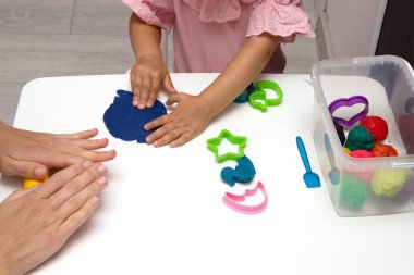 Beyaz bir masanın üzerinde renkli plastikçiden bebek heykelleri. Küçük bir çocuğun eli renkli plastiğin parçalarını sıkıyor. Anne kızıyla eğitim oyunları oynuyor.