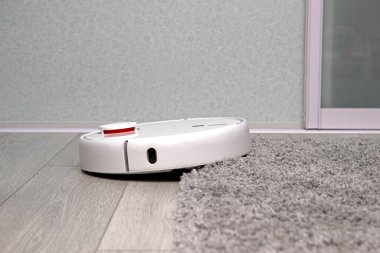 Beyaz robot elektrikli süpürge halıda sürüyor. Robot elektrik süpürgesiyle engelleri aşmak. Modern kablosuz cihazlarla ev temizliği yapmak için ideal. akıllı ev kavramı.