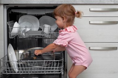 Sevimli sarışın bebek mutfakta yardım ediyor bulaşık makinesinden tabakları çıkarıyor. Çocuk ev işlerine yardım ediyor.