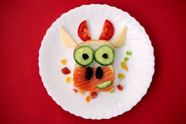 Yenebilir 2021 boğa ineği sembollü komik sandviç kızarmış ekmek, kırmızı balık, salatalık, domates, peynir ve kuru üzümden yapılır. Çocuklar için kahvaltı fikri. Yeni yıl yemeğinin üstü açılır. yemek sanatı