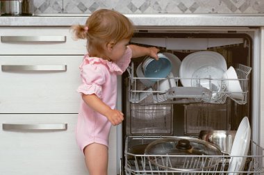 Sevimli sarışın bebek mutfakta yardım ediyor bulaşık makinesinden tabakları çıkarıyor. Çocuk ev işlerine yardım ediyor.