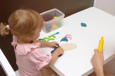 Beyaz bir masanın üzerinde renkli plastikçiden bebek heykelleri. Küçük bir çocuğun eli renkli plastiğin parçalarını sıkıyor. Anne kızıyla eğitim oyunları oynuyor.