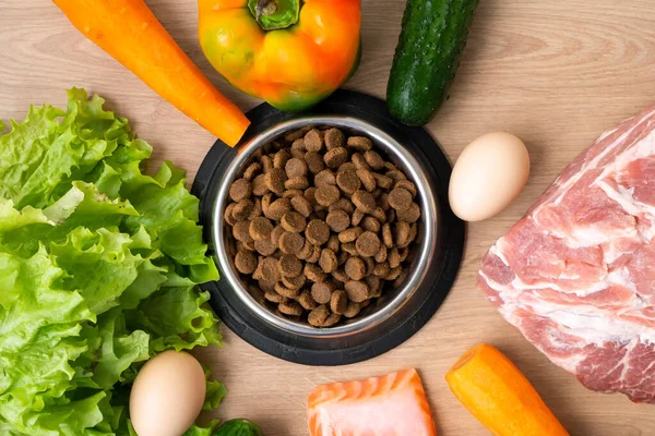 Dry pet dog food with natural ingredients. Raw meat, fish, vegetables, eggs and salad near bowl with dry pet feed on wooden background. concept of correct balanced nutrition for pet, flatly