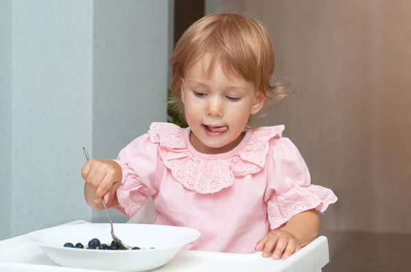 Menina pequena comendo sua aveia de mingau de manhã com mirtilos ela mesma, sentada em cadeira de bebê na cozinha — Fotografia de Stock