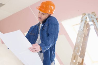 Eller planı tutarak ve onun cep telefonu, merdiven dururken konuşurken orta yaşlı inşaat işçisi portresi. 