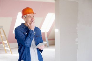 Orta yaşlı inşaat işçisi inşaat sitesinde ayakta ve dijital tablet kullanarak düşünceli arıyorum portresi. 