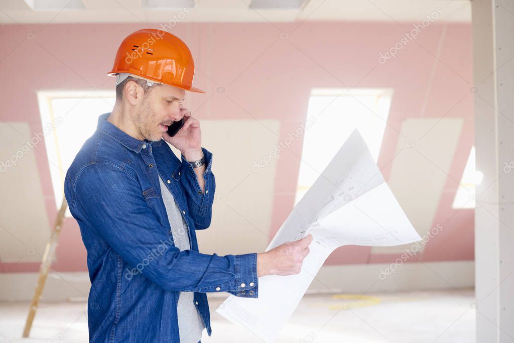 Middle aged construction worker holding plan in his hand while using mobile phone and talking with somebody.