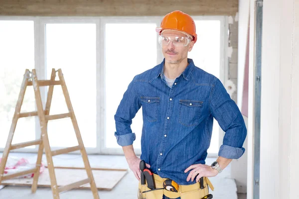 Portrait of thinking handyman wearing safety helmet and protective eye goggles while standing with hands on his hip at the construction site.
